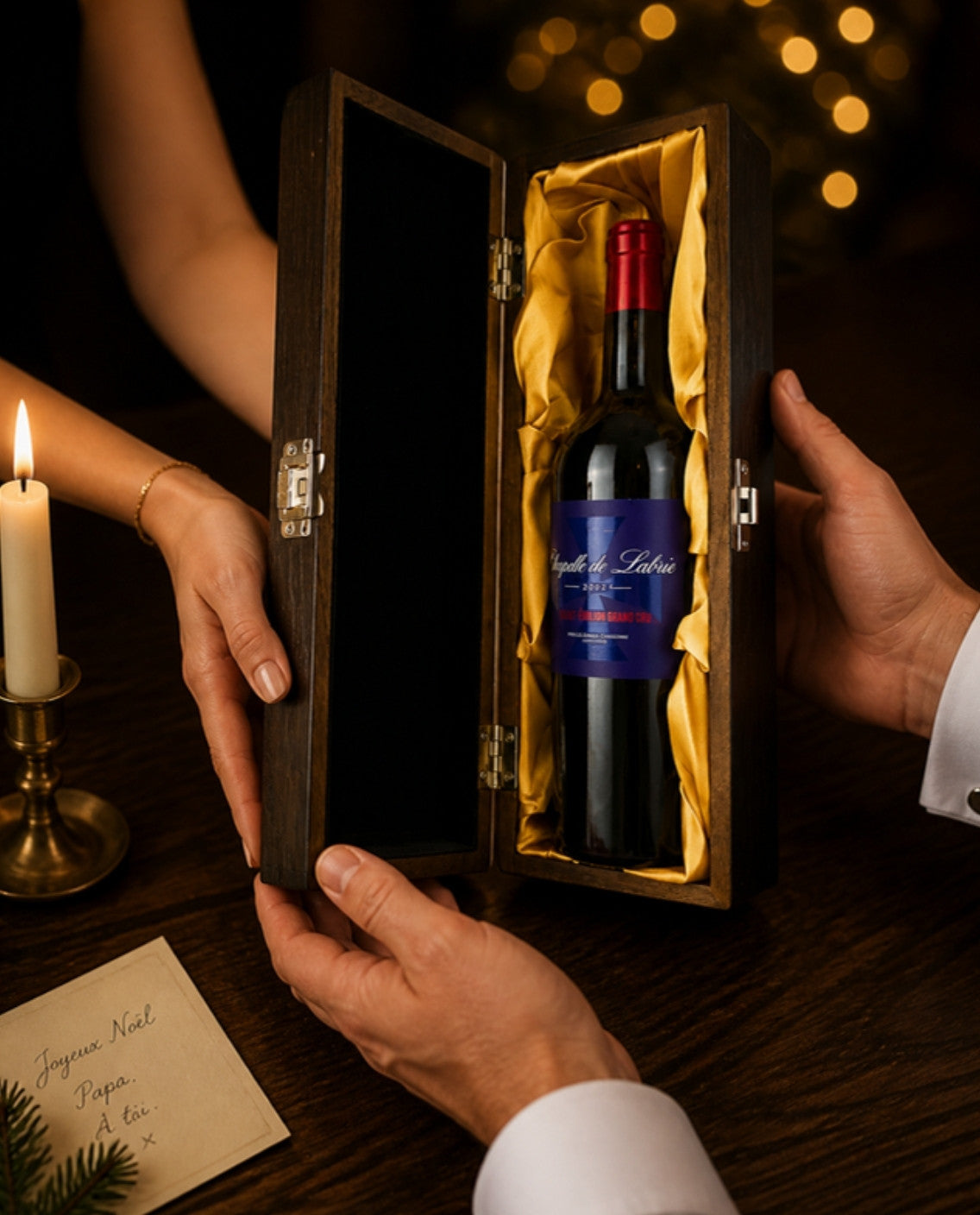Hands passing the Chapelle de Labrie 2021 Saint-Émilion Grand Cru in its elm wood gift box with gold lining across a candlelit table at Christmas