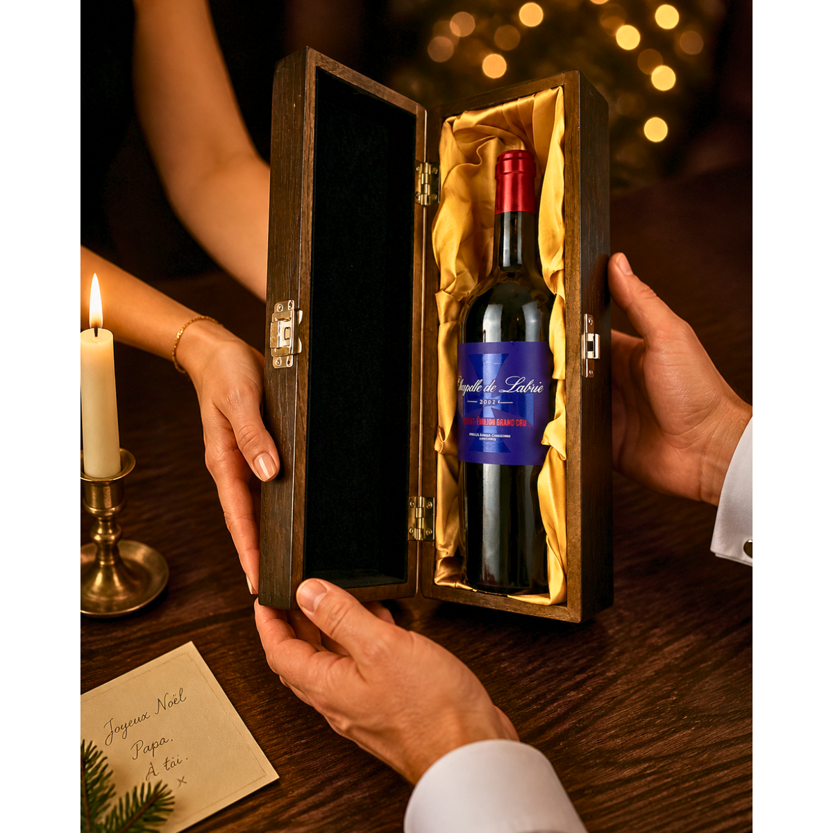 Hands passing the Chapelle de Labrie 2021 Saint-Émilion Grand Cru in its elm wood gift box with gold lining across a candlelit table at Christmas